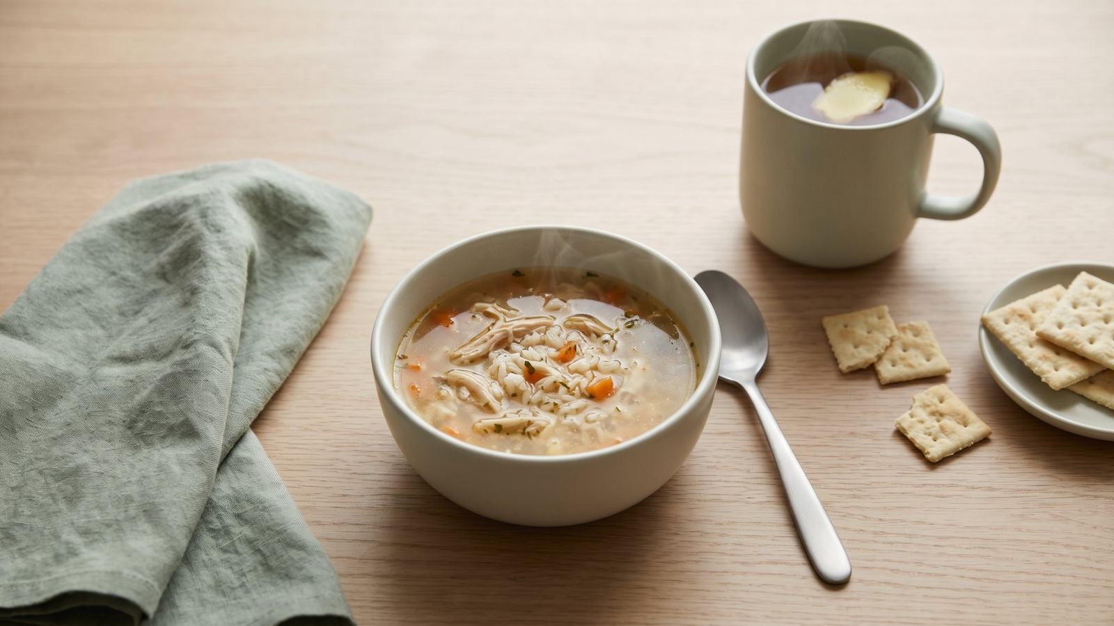 A small bowl of chicken and rice soup, a mug of ginger tea and a few plain crackers on a light table