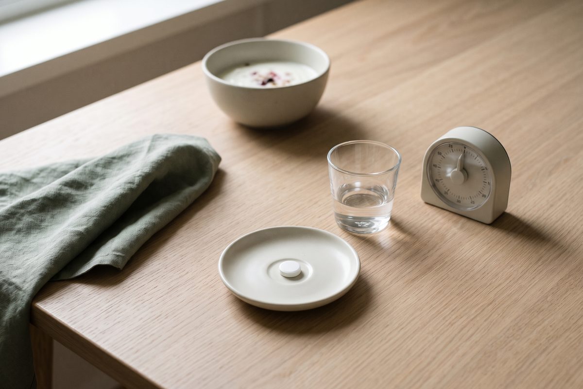 A plain tablet on a saucer, a small glass of water and a kitchen timer, with a bowl of yogurt in the background